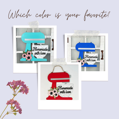 The Kitchen Mixer Wall Hanger DIY Home Decor Craft Kit by Janet's Craft Corner features three paper cutouts of stand mixers in blue, teal, and red with "Homemade with Love" signs. Adorned with pink flowers on a polaroid-style backdrop, it asks "Which color is your favorite?" to enhance kitchen charm.