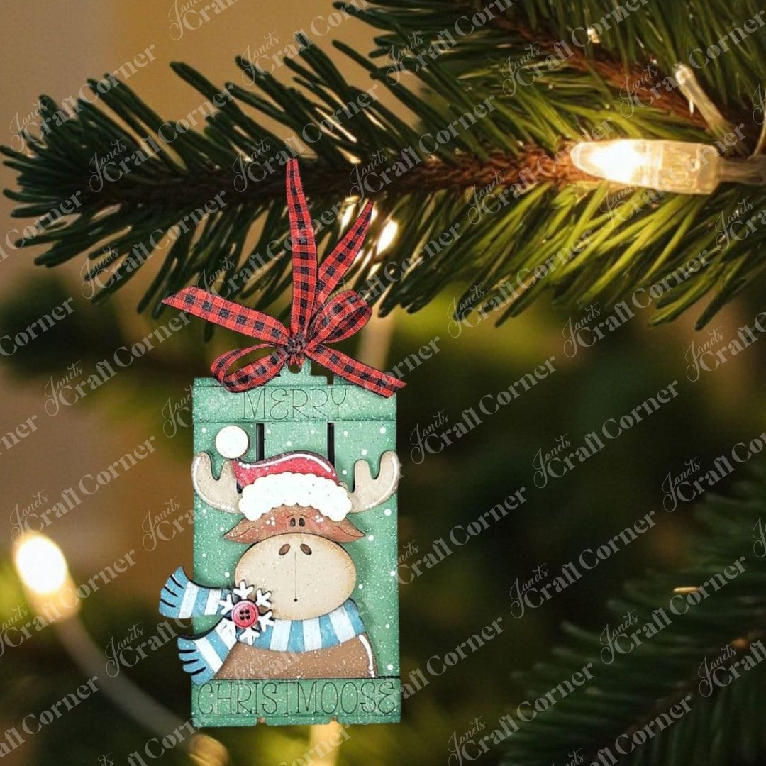 A Christmas tree branch with a string of lights features a Forest Friends Ornaments DIY craft kit moose from Janet's Craft Corner, wearing a Santa hat and scarf, with "Merry Christmoose" written on it—ideal for animal ornament lovers!.
