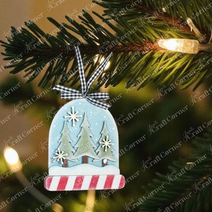 A snowy scene ornament with pine trees, snowflakes, and cute animals from Janet's Craft Corner’s Forest Friends Ornaments DIY craft kits (choice of 5) hangs by a black and white ribbon among glowing lights.