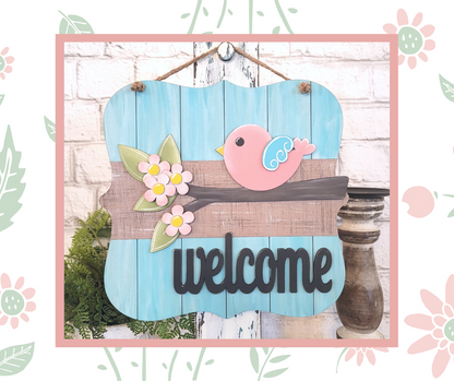 The Bird Welcome Sign from Janet's Craft Corner is a DIY seasonal home decor craft kit featuring a pink bird on a floral-branch against a blue wood backdrop, with "welcome" in bold black letters, perfect for adding charm to your entryway.