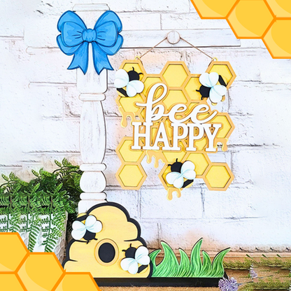 The "Bee Happy Street Post Shelf Sitter" by Janet's Craft Corner adds charm with hexagonal honeycomb shapes, the phrase "bee happy," a yellow beehive in green grass, and small bees against a white brick backdrop.