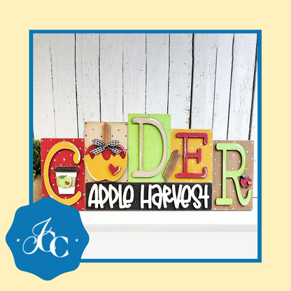 The Cider Word Block Shelf Sitter from Janet's Craft Corner is a DIY seasonal home decor craft kit. Colorful letters spell "CIDER" with apple motifs and patterns over "APPLE HARVEST," all on a rustic white wooden panel. A blue logo with stylized initials adorns the bottom left corner of this charming piece.