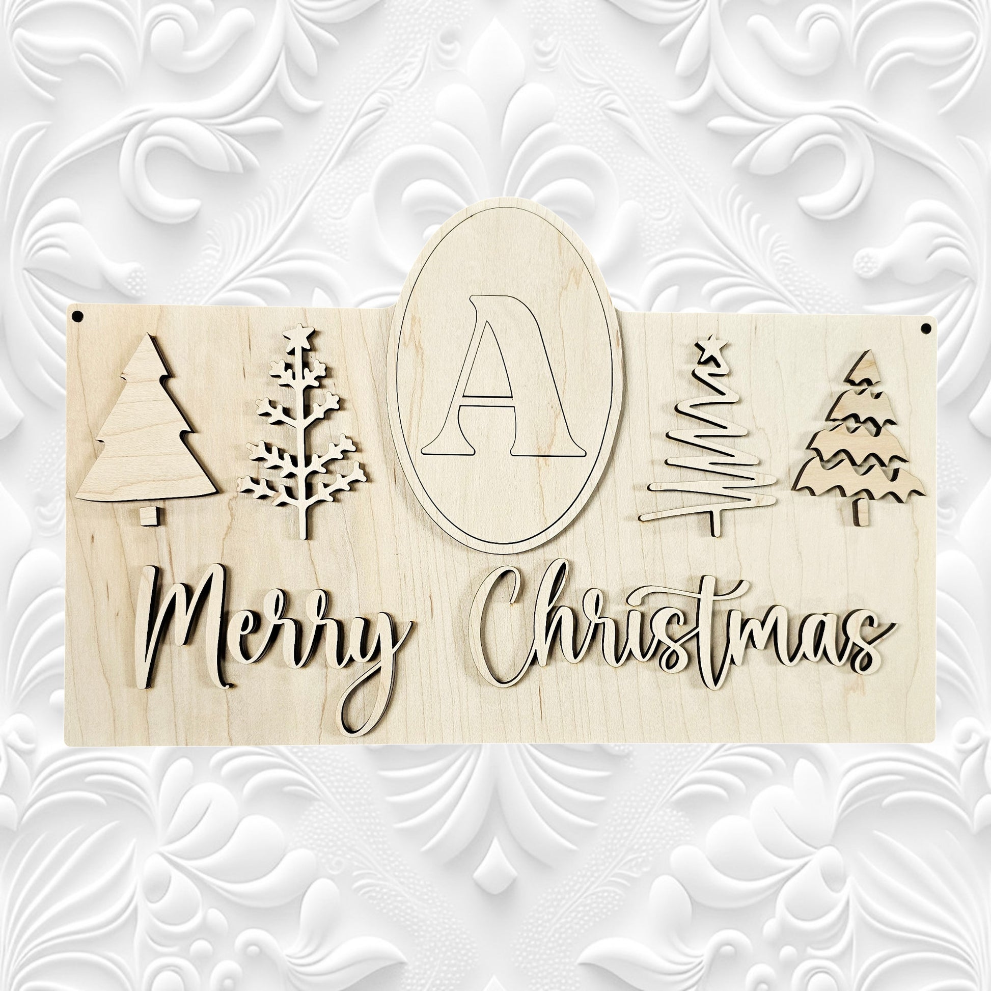 The Monogram Trees Wall Hanger by Janet's Craft Corner is a DIY home decor craft kit featuring wooden Christmas trees and the phrase "Merry Christmas." It includes an oval with the letter "A" at the top, set against a background with a decorative white embossed pattern, making it perfect for festive displays.