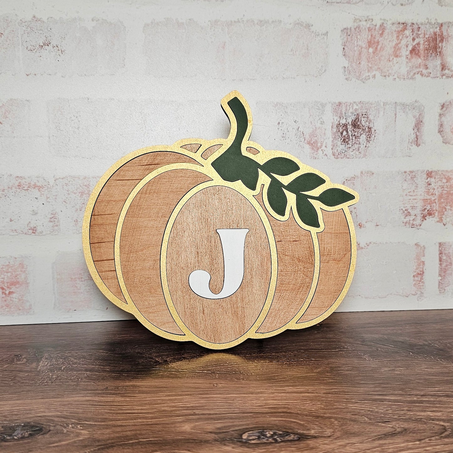 The Monogram Pumpkin Shelf Sitter, part of Janet's Craft Corner's DIY home decor craft kit, features a captivating wooden pumpkin design with the letter "J" at its center and a green leaf on top. This delightful personalized decoration sits gracefully against a light brick wall on a wooden surface, bringing an inviting ambiance to any room.