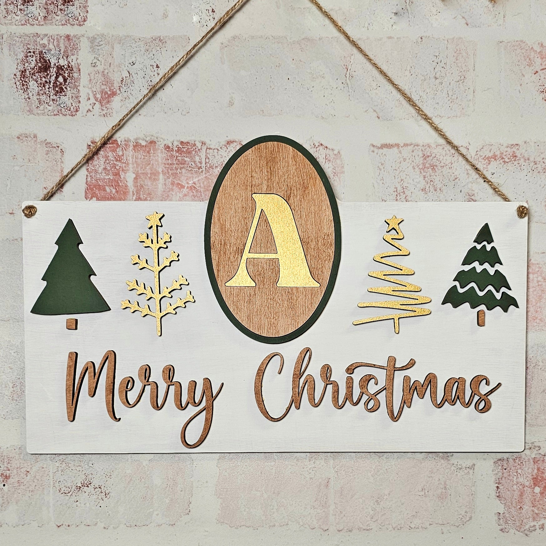 A delightful DIY craft kit from Janet's Craft Corner, the Monogram Trees Wall Hanger features a wooden wall sign showcasing a central "A" monogram surrounded by tree designs. Elegantly scripted with "Merry Christmas" below, this charming piece of home décor hangs from a rope against a brick wall, infusing any space with warmth and festive cheer.