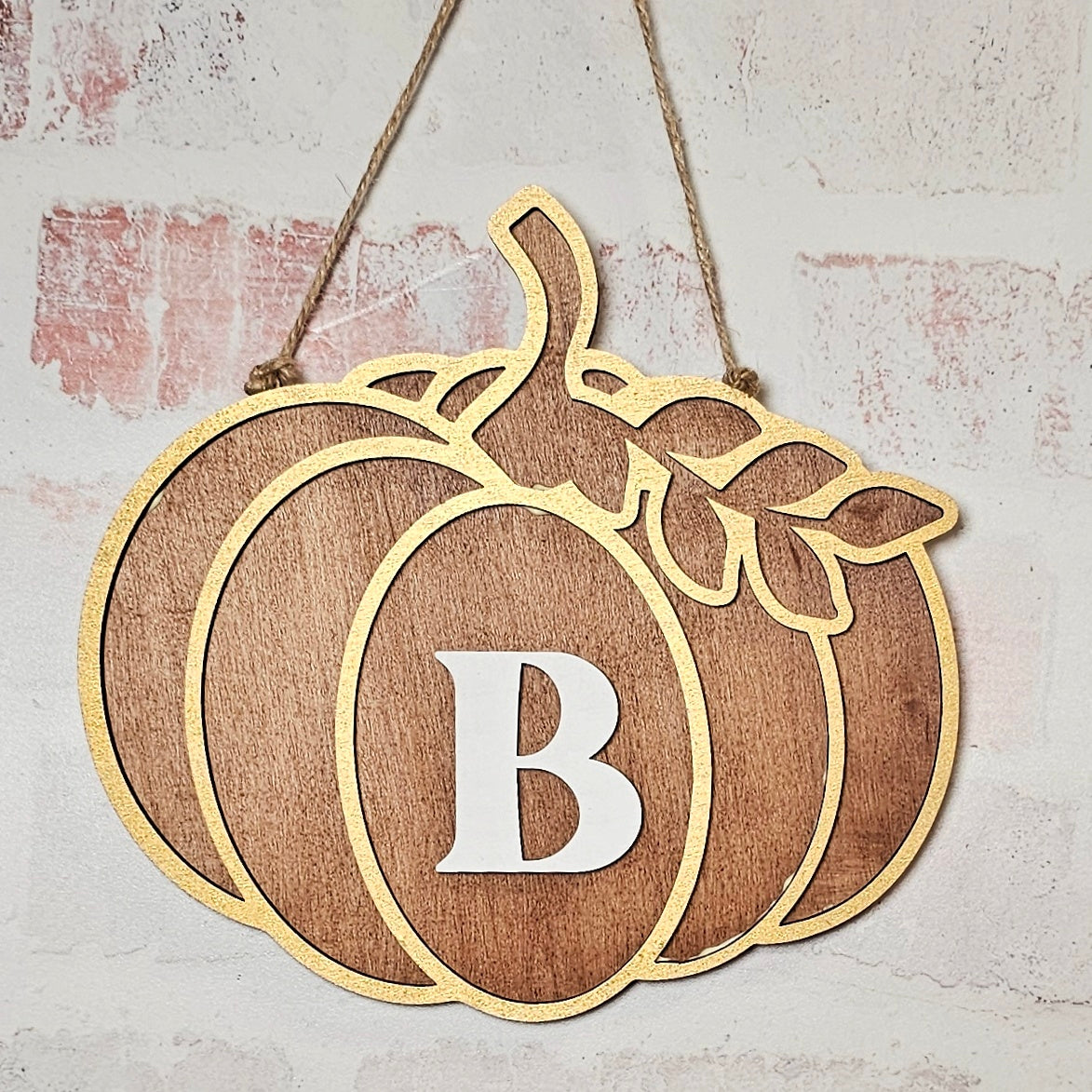 Janet's Craft Corner offers a Monogram Pumpkin Wall Hanger DIY home decor craft kit, featuring a delightful hand-painted wooden decoration with a letter "B" outlined in gold glitter. The wall hanger, suspended effortlessly against a brick backdrop with a twine loop, adds an elegant touch to your DIY home decor projects.