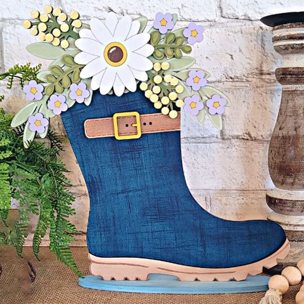 The Floral Rainboot Shelf Sitter by Janet's Craft Corner is a decorative piece featuring colorful flowers like daisies and purple blossoms spilling from its top. It includes a yellow buckle and rests on a burlap surface, making it ideal for home décor. In the background, there's a plant against a white brick wall.