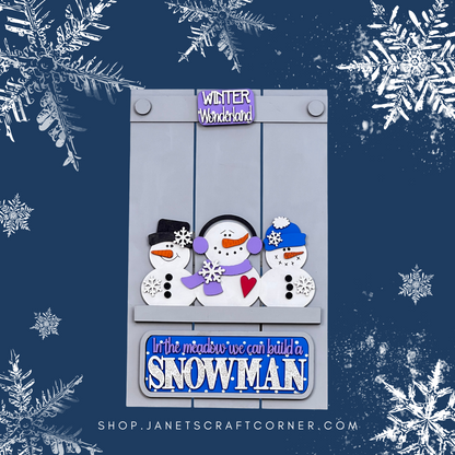 The Snowman Interchangeable Insert DIY kit by Janet's Craft Corner features a decorative winter sign with three snowmen in various hats and scarves, surrounded by snowflakes. The hand-painted kit includes "Winter Wonderland" and "In the meadow we can build a snowman." Shop link available below.