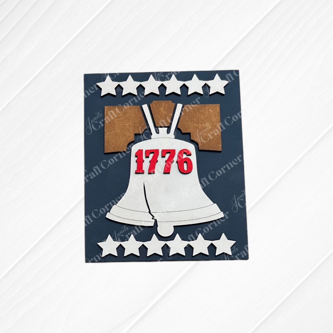Janet's Craft Corner's Patriotic Farmhouse Signs DIY kit features a Liberty Bell with "1776" in red, seven white stars, and a white wood pattern—ideal for spring tiered tray decor or creative farmhouse sign projects. Set of 1.