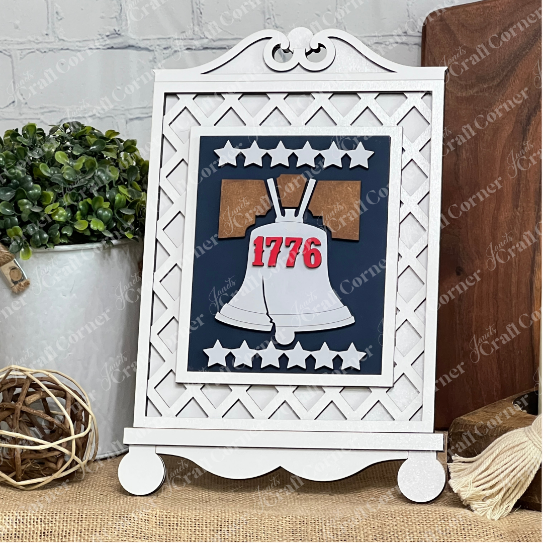 The Cottage Frame for Switchable Farmhouse Signs by Janet's Craft Corner displays the Liberty Bell with "1776" in red and white stars, perfect for seasonal decor. Green plants and a rustic background add to this charming DIY craft kit scene.