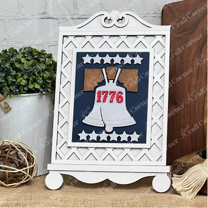 The Cottage Frame for Switchable Farmhouse Signs by Janet's Craft Corner displays the Liberty Bell with "1776" in red and white stars, perfect for seasonal decor. Green plants and a rustic background add to this charming DIY craft kit scene.