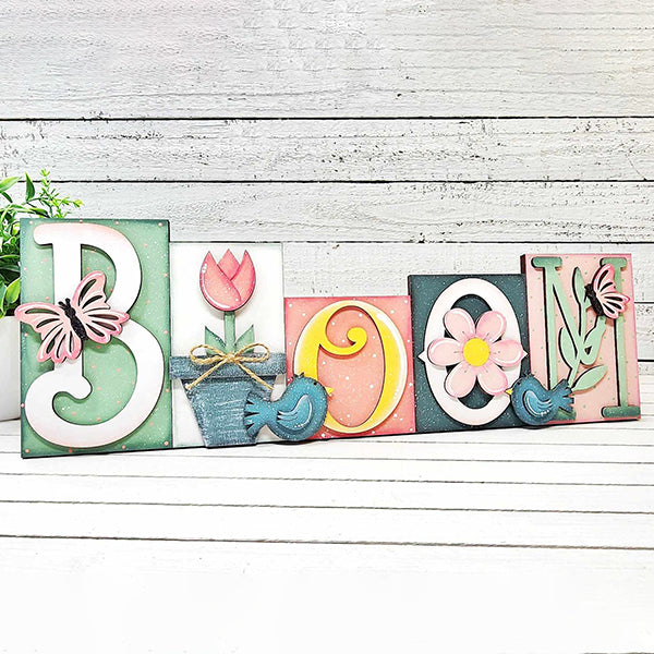 The Bloom Word Block by Janet's Craft Corner spells "BLOOM" with charming floral and butterfly motifs, including a tulip, daisy, and pastel-colored butterflies. This piece is perfect for enhancing home décor against a white wooden wall.