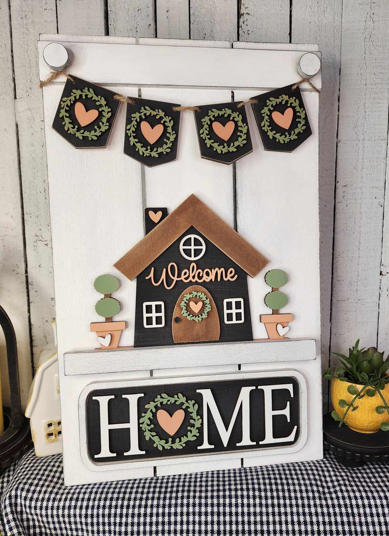 The Welcome Home Interchangeable Insert by Janet's Craft Corner is a DIY home decor kit featuring a hand-painted decorative wooden sign. The sign showcases a house with "Welcome" inscribed, pennants with heart designs above, and the word "HOME" embellished with a heart and wreath motif below. It comes thoughtfully arranged on a checked cloth near a plant, making it perfect for customizable home decor.