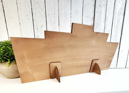 The Cider Word Block Shelf Sitter from Janet's Craft Corner, featuring a geometric stepped design with two arches, stands on a white surface. Perfect as home décor, it's set against whitewashed wooden planks with a nearby leafy plant in view.