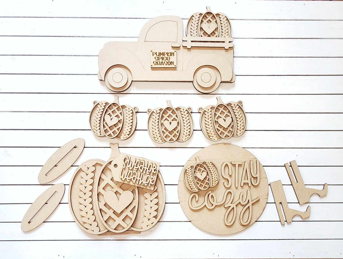 Janet's Craft Corner presents the Sweater Weather Decor Set, a DIY home decor craft kit that includes wooden cutouts on a white background featuring a truck with "Pumpkin Spice Season," a plaque with "Sweater Weather," and a "Stay Cozy" sign, complemented by pumpkins and boots. This set is perfect for autumn crafts and home decor projects.