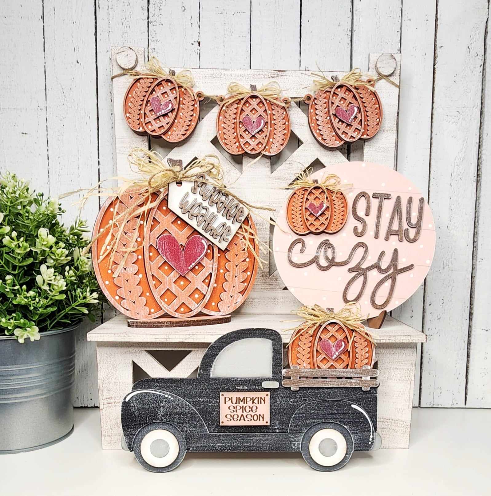 A cozy autumn display showcases delightful home decor with wooden accents. A black truck labeled "Pumpkin Spice Season" is loaded with pumpkins, while a large pumpkin sits next to a "Stay Cozy" sign, all enhanced by the enchanting Sweater Weather Decor Set from Janet's Craft Corner. Completing the scene, a small plant adds the perfect touch.