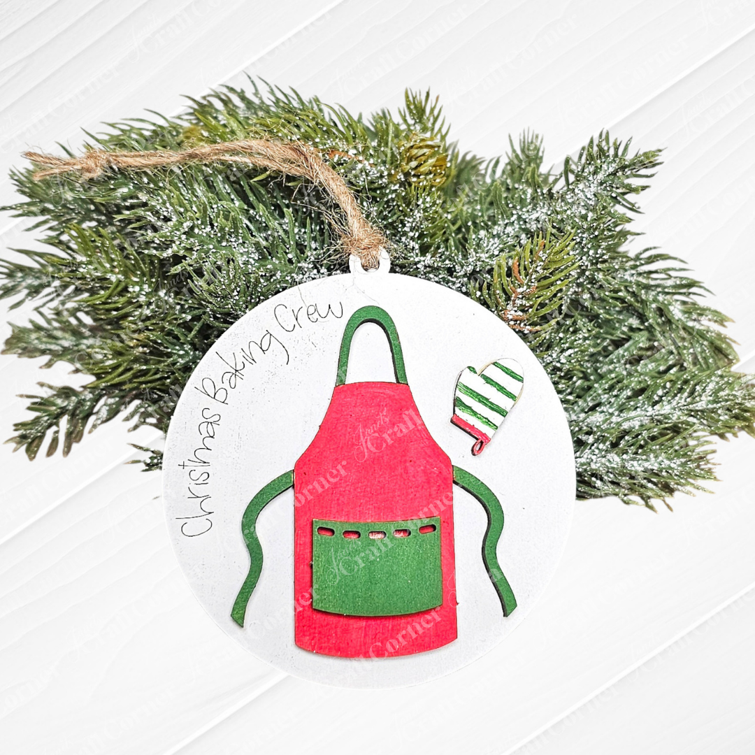 This delightful addition to your home décor is part of the "Baking Ornaments - Set of 2" from Janet's Craft Corner. It includes a round Christmas ornament showcasing a red apron with green pockets and a striped green and white oven mitt, with the phrase "Christmas Baking Crew." Nestled against frosted pine branches, this piece is perfect for enhancing your festive baking decorations.