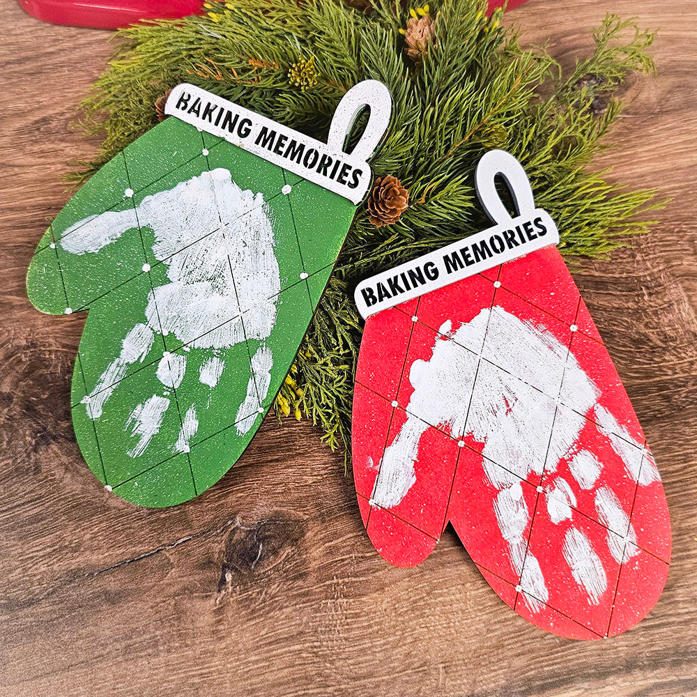 Two delightful Christmas-themed ornaments from the "Baking Memories Kids Pop Out Kit" by Janet's Craft Corner, shaped like oven mitts in green and red with white handprints, bring a festive flair to your home décor. Labeled "Baking Memories" on the cuff, they evoke warmth against a wooden backdrop adorned with greenery.