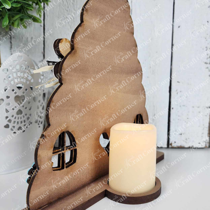 Beside a lit LED candle on a white wooden surface, Janet's Craft Corner Honey Bee Mini Shelf Sitter Set showcases a wooden triangular-shaped fairy house with window cutouts, perfect for bee-lovers desiring whimsical DIY decor with a natural finish and soft glow.
