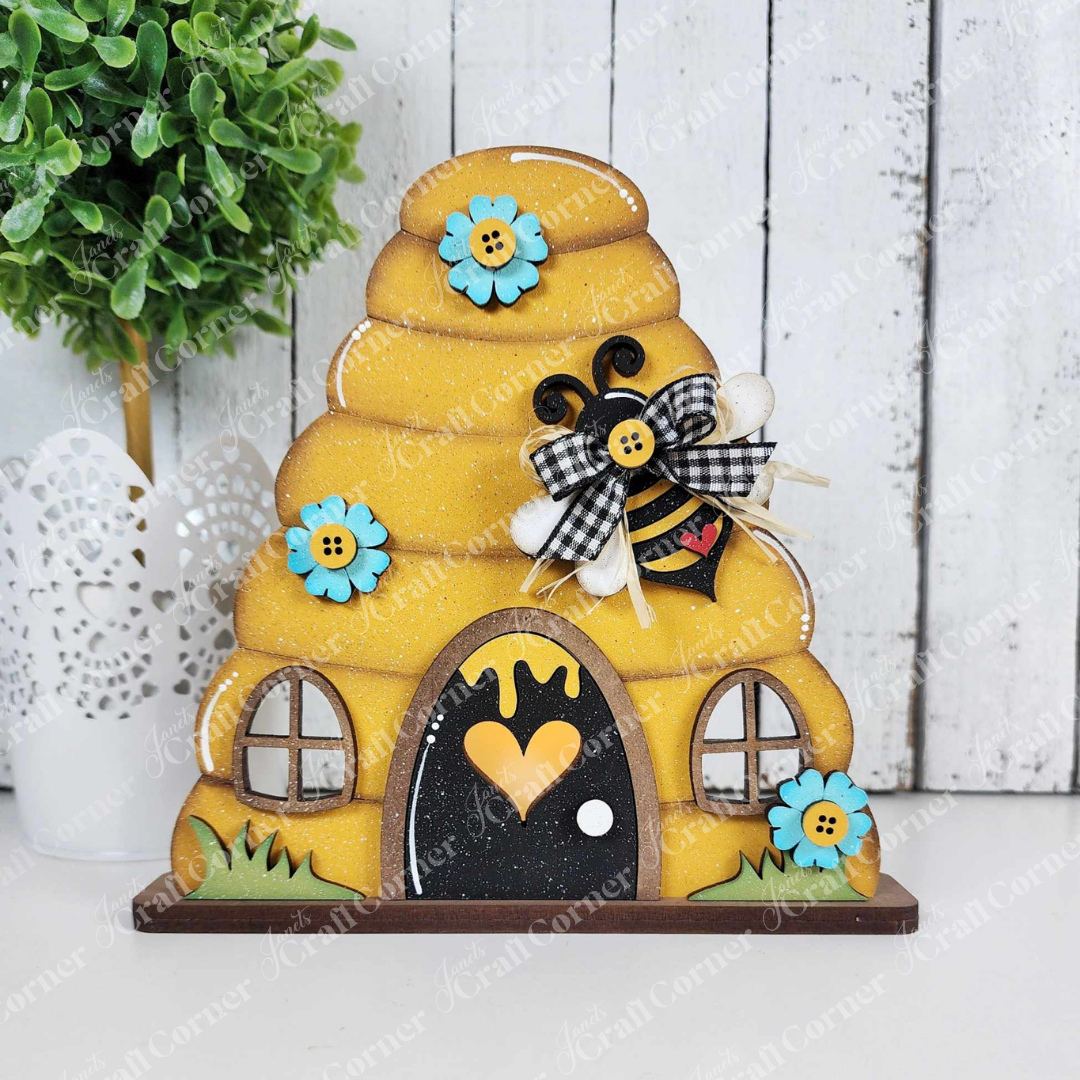 This delightful bee-themed wooden beehive, featuring a heart-shaped entrance, two windows, blue flowers, and a checkered-bow bee against a white textured background with a green potted plant, is perfect for any bee-lover's collection. Part of Janet's Craft Corner's Honey Bee Mini Shelf Sitter Set.