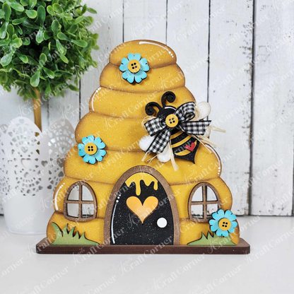 This delightful bee-themed wooden beehive, featuring a heart-shaped entrance, two windows, blue flowers, and a checkered-bow bee against a white textured background with a green potted plant, is perfect for any bee-lover's collection. Part of Janet's Craft Corner's Honey Bee Mini Shelf Sitter Set.