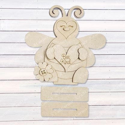 Janet's Craft Corner presents the Honey Bee Mini Shelf Sitter Set—a wooden cutout of a smiling bee holding a heart with "Bee Kind," perfect for bee-lovers. This DIY seasonal home decor kit includes three blank slots and rests on white wooden planks, ready for personal touches.