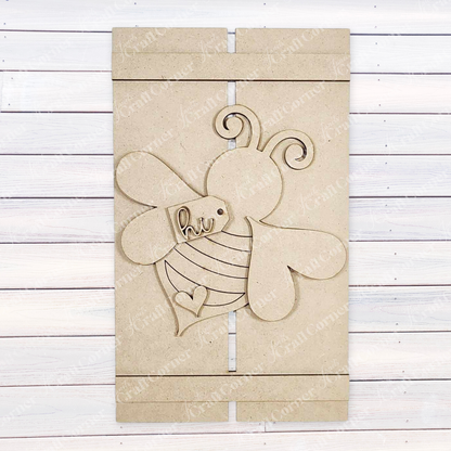 The Bee Pallet Sign Shelf Leaner from Janet's Craft Corner is a DIY craft kit featuring a wooden panel with a cutout bee adorned with curly antennae, heart-shaped wings, and an engraving of "hi" and a small heart, creating charming home decor against its white wooden background.