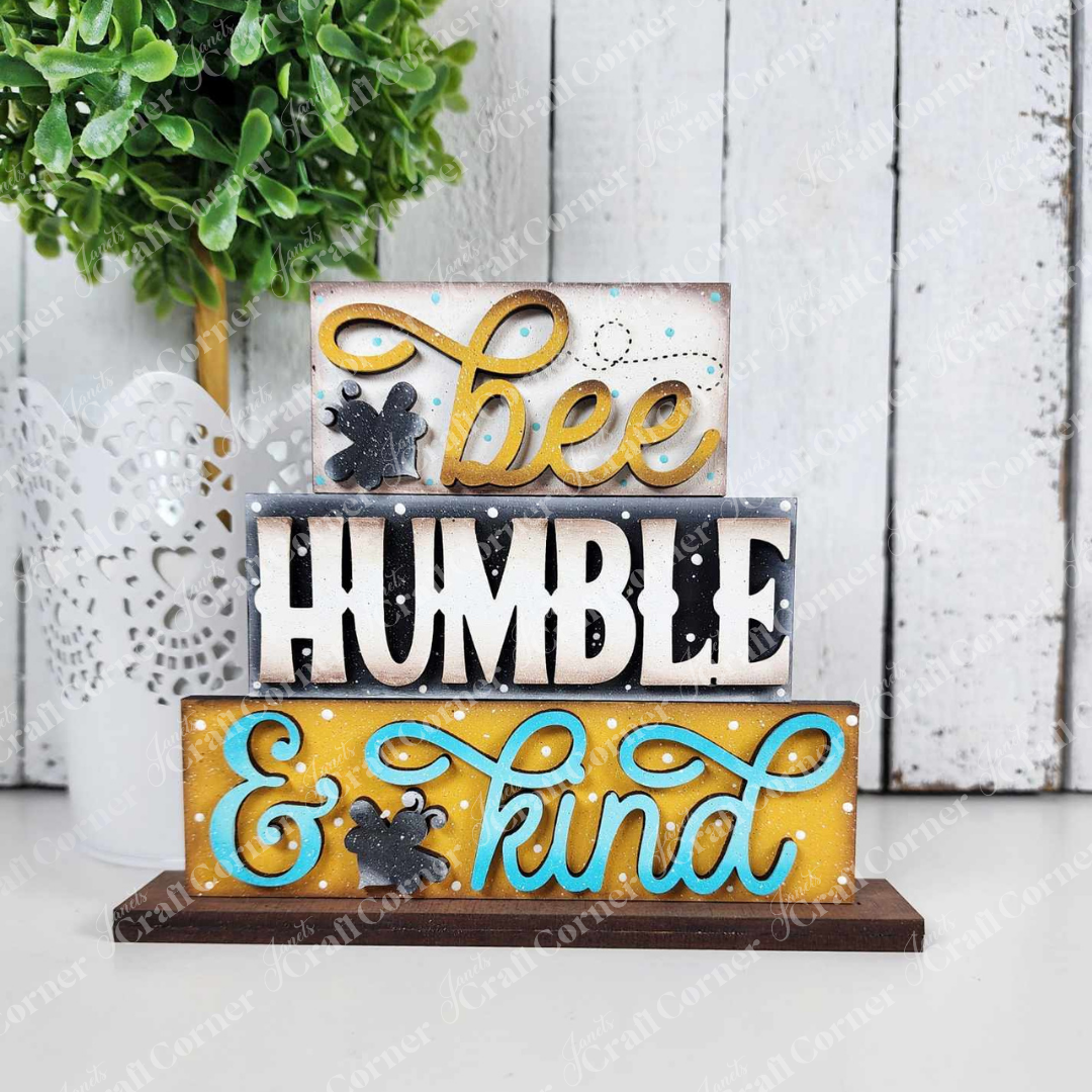 The Honey Bee Mini Shelf Sitter Set from Janet's Craft Corner offers three stacked wooden signs with bee and honeycomb designs. Featuring the words "bee" and "kind" in yellow and blue with decorative swirls, it includes a white wooden wall backdrop and a potted plant, ideal for bee-lovers.