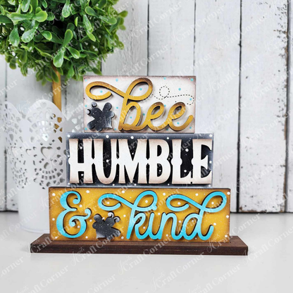 The Honey Bee Mini Shelf Sitter Set from Janet's Craft Corner offers three stacked wooden signs with bee and honeycomb designs. Featuring the words "bee" and "kind" in yellow and blue with decorative swirls, it includes a white wooden wall backdrop and a potted plant, ideal for bee-lovers.