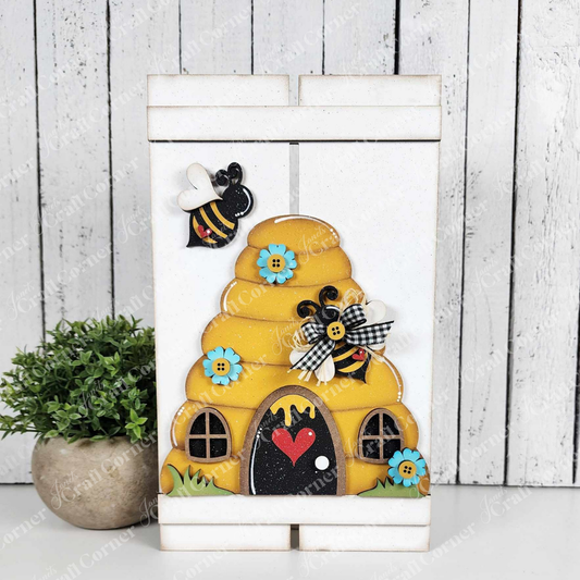 Craft a delightful home decor piece with Janet's Craft Corner's Beehive Pallet Sign Shelf Leaner DIY kit. This art features bee embellishments, blue flowers, a heart detail at the entrance, and a small potted plant against a wooden panel backdrop.