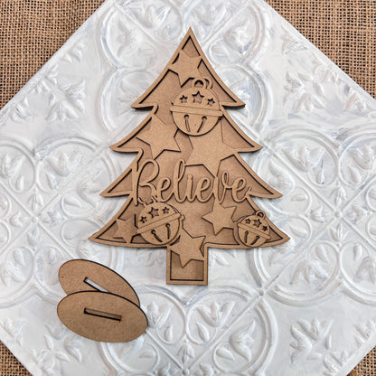 The Standing Christmas Tree Shelf Sitters by Janet's Craft Corner, featuring "Believe" carved with star and bell designs, make the perfect addition to your holiday decor. Placed on a textured white and brown background next to two small wooden pieces, they're ideal for a charming shelf display.