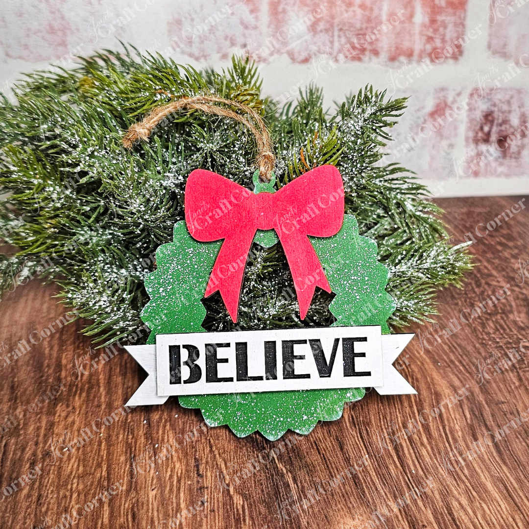 A charming, hand-painted green wreath from Janet's Craft Corner features a red bow and the word "BELIEVE" on a white banner. Glitter accents enhance its festive appeal as it hangs from twine against pine branches and a brick wall, beautifully complementing your Holiday Village Ornament Set - Early Access.