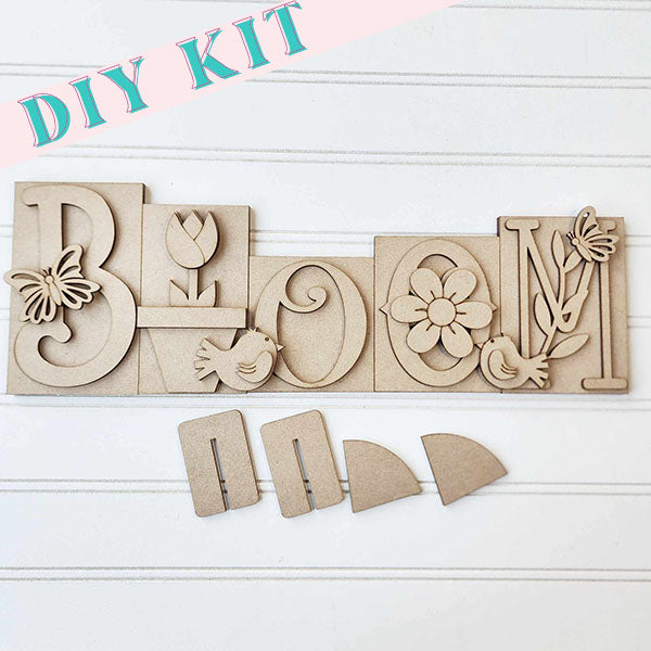 Introducing the Bloom Word Block by Janet's Craft Corner: This DIY kit includes charming wooden letters spelling "Bloom," each embellished with intricate floral and bird motifs. Ideal for home décor, these letters are set against a white, textured background, accompanied by additional wooden pieces below to elevate any living space.