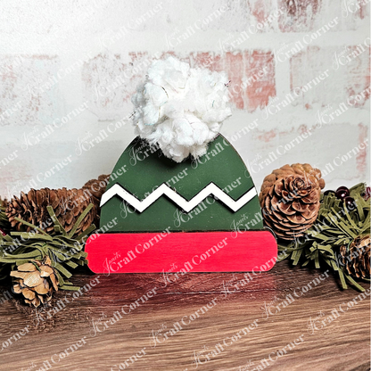 The Build a Beanie Pop-Out by Janet's Craft Corner features a festive wooden elf hat with a green top, white zigzag trim, red brim, and white pom-pom. Hand-painted and set against pinecones and greenery, it's perfect for adding charm to your home décor.