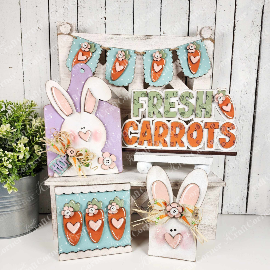 The Bunnies & Carrots Decor Set from Janet's Craft Corner includes bunny cutouts, a "Fresh Carrots" sign, and carrot garlands. These pastel pieces are set against a rustic white background with a small plant. Personalize your space with this hand-painted DIY craft kit.