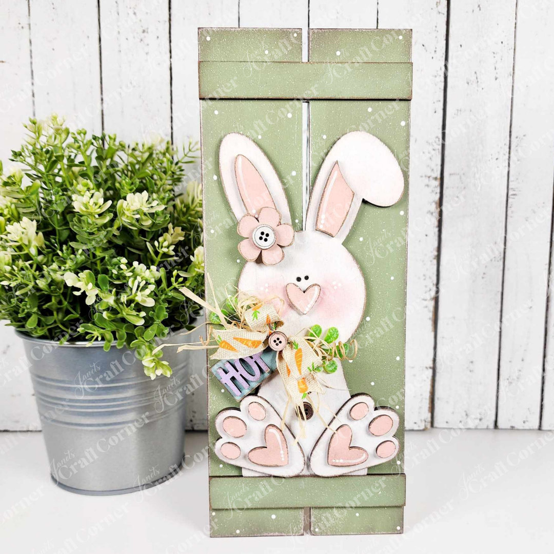 Janet's Craft Corner's Bunny Table Top Pallet craft kit features a wooden Easter bunny with a pink nose and button eyes, holding a "HOP" sign beside a green potted plant. Perfect as a shelf leaner or carrot décor piece, it adds charm with its light-colored background.
