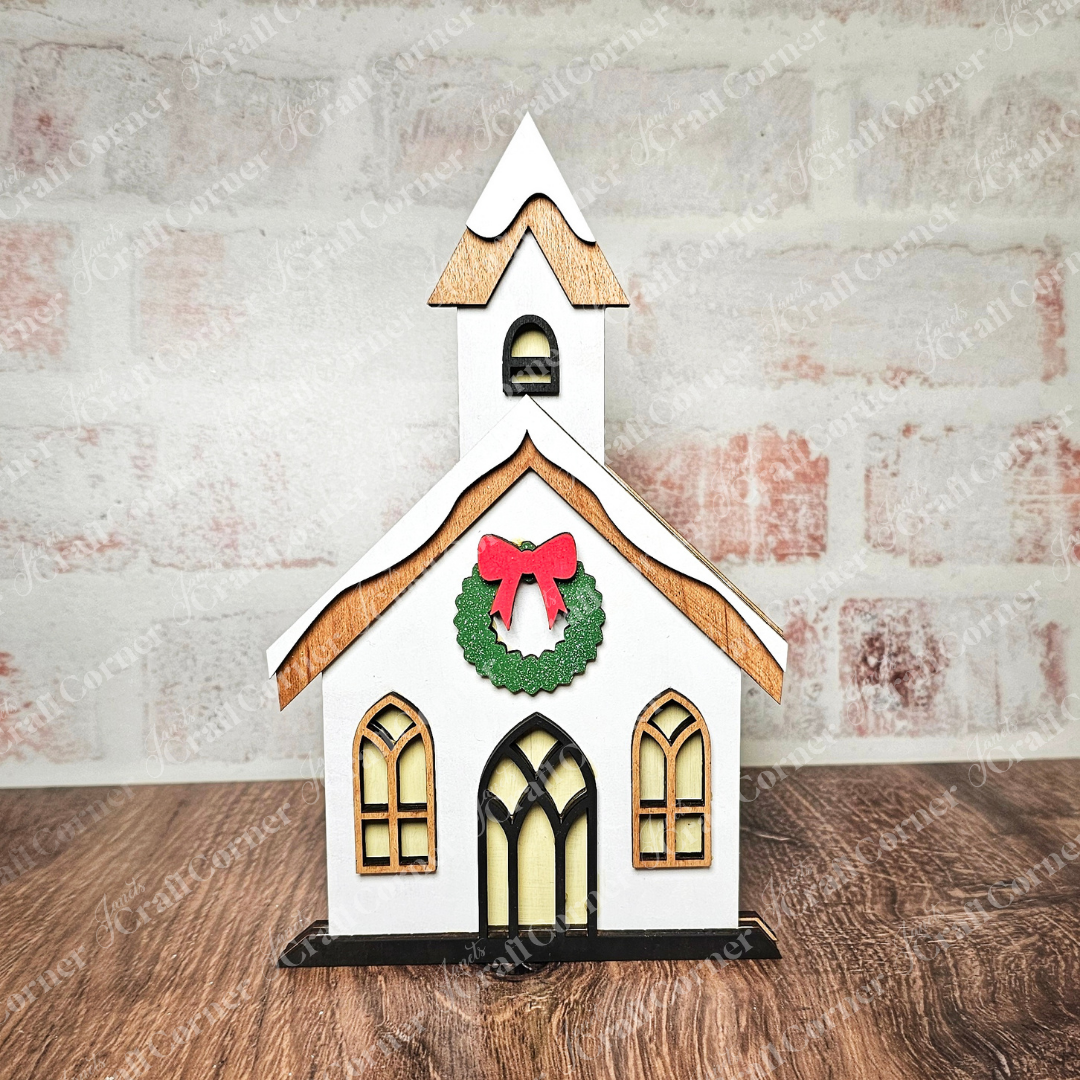 This hand-painted Christmas village shelf sitter from Janet's Craft Corner is a charming decorative wooden model of a white church with a brown roof, adorned with a green wreath and red bow above the door. It's perfect for home décor, resting beautifully on any wooden surface against a brick wall.