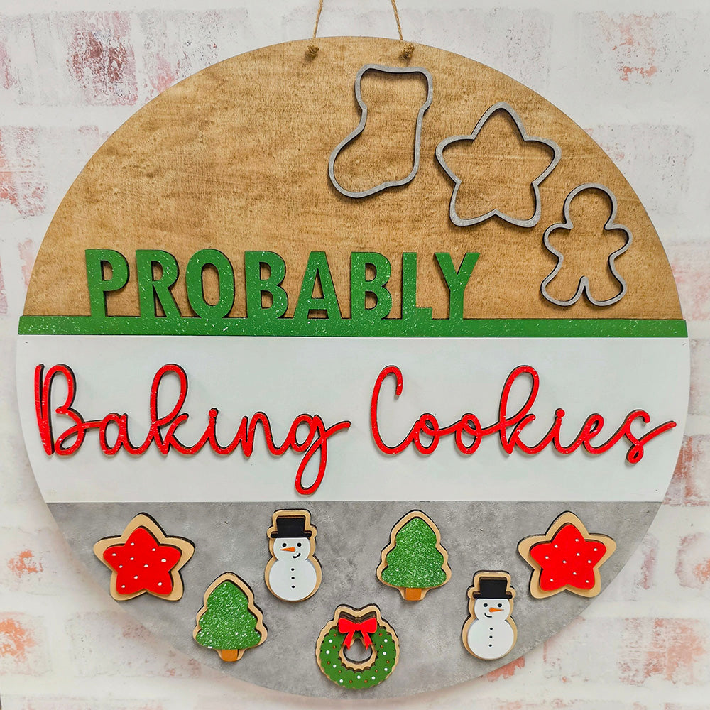 The "Probably Baking Cookies Door Hanger - DIY Home Decor Kit" from Janet's Craft Corner is a round wooden sign that features cookie cutters and displays the phrase "Probably Baking Cookies" in festive green and red colors. This hand-painted craft kit is adorned with charming cookie shapes such as stars, snowmen, and trees at the bottom, bringing a personalized touch to your DIY home decor.