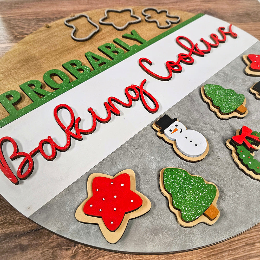 Introducing the Probably Baking Cookies Door Hanger - DIY Home Decor Kit by Janet's Craft Corner: This delightful set features a wooden board with cookie cutters arranged to spell "Probably" above and "Baking Cookies" below. Complementing the text are charming cookies in the shapes of a snowman, wreath, Christmas tree, and star, each beautifully decorated with vibrant icing to add a personal touch to your home decor.