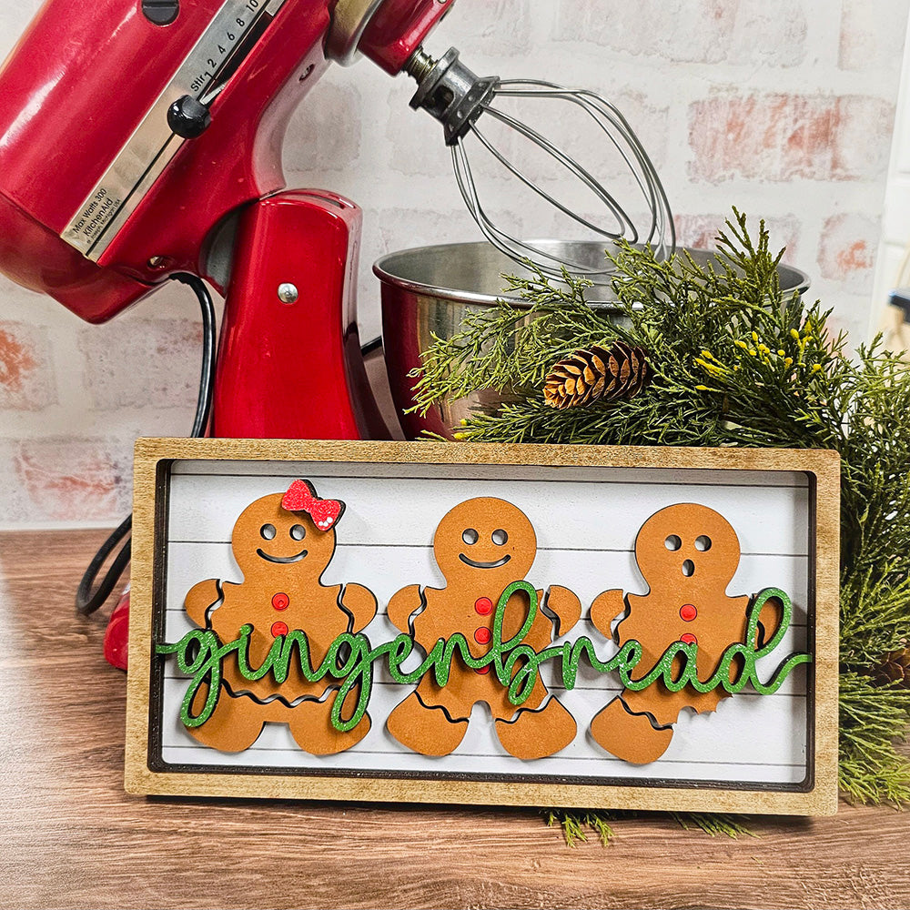 A wooden sign featuring three gingerbread figures, each decorated with red buttons and one wearing a red bow tie, showcases the word "gingerbread" in green script. A red mixer and pine branches provide a festive backdrop. This delightful piece is ideal for crafting enthusiasts interested in the Gingerbread Chunky Shelf Sitter DIY Home Decor Kit from Janet's Craft Corner.