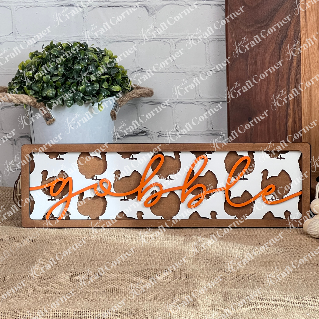 Introducing Janet's Craft Corner's "Gobble Turkey Chunky Shelf Sitter," a charming DIY home decor craft kit. This kit includes a wooden sign showcasing the word "gobble" in vibrant orange cursive against a white background adorned with brown turkey silhouettes. Enhance your cozy home ambiance by placing it alongside a potted plant, set against a white brick wall, and displayed on a burlap-covered surface scattered with autumn leaves.