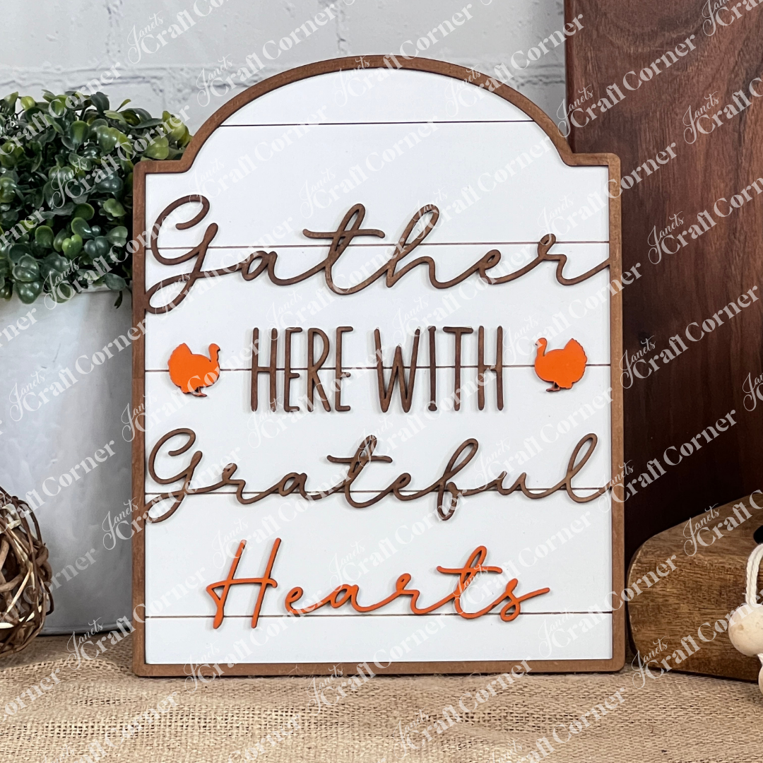 The Grateful Hearts Shelf Leaner from Janet's Craft Corner, adorned with the phrase "Gather Here with Grateful Hearts," graces the shelf next to a small plant and basket. With its orange accents and wooden frame, it brings a cozy, rustic charm to your home décor.