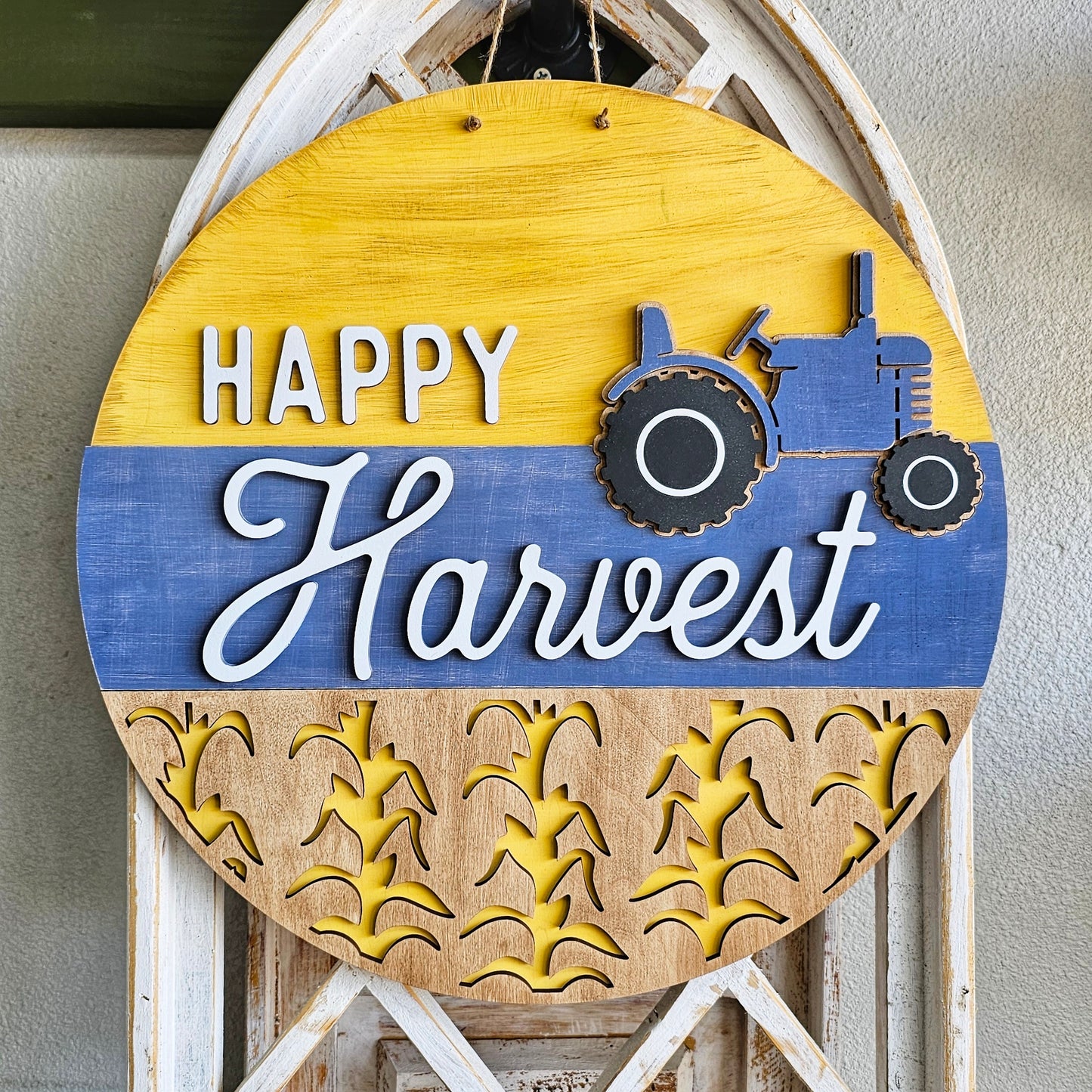 Introducing the Happy Harvest Door Hanger DIY home decor kit from Janet's Craft Corner. This round wooden sign adds a charming touch to any space, with "Happy Harvest" in white letters. It features a yellow top with a blue tractor and a bottom adorned with cut-out wheat stalks on a blue background, all beautifully hand-painted.