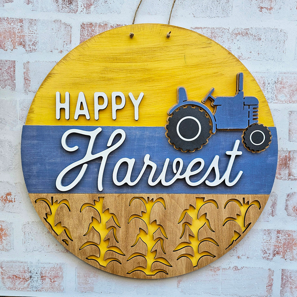 This delightful DIY home decor kit from Janet's Craft Corner, the Happy Harvest Door Hanger, includes a round wooden sign with "Happy Harvest" prominently displayed above a blue tractor. The sign is artistically divided into vibrant yellow and blue sections and is adorned with wheat stalks at the bottom, making it an ideal addition to bring rustic charm to your home decor against a brick wall.