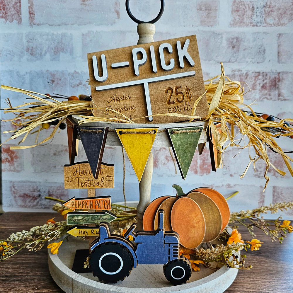 A charming display for the fall season can be created with Janet's Craft Corner's Harvest Festival Decor Set - 5 pieces. The set includes a delightful "U-PICK" sign offering apples and pumpkins for 25 cents per pound, accompanied by a small wooden tractor, a pumpkin, and colorful pennants. Perfectly rustic against a backdrop of straw and autumn leaves, it's an ideal choice for Harvest Festival Decor.