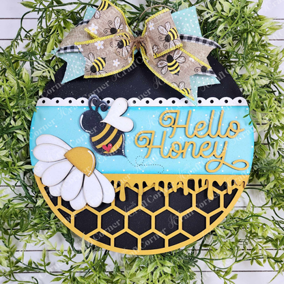The Hello Honey Wall Hanger from Janet's Craft Corner features a bee, daisies, and honeycomb design with "Hello Honey" text. An aqua blue and black background is surrounded by green foliage with a burlap bow adorned with bee motifs on top, adding charm to any home decor.
