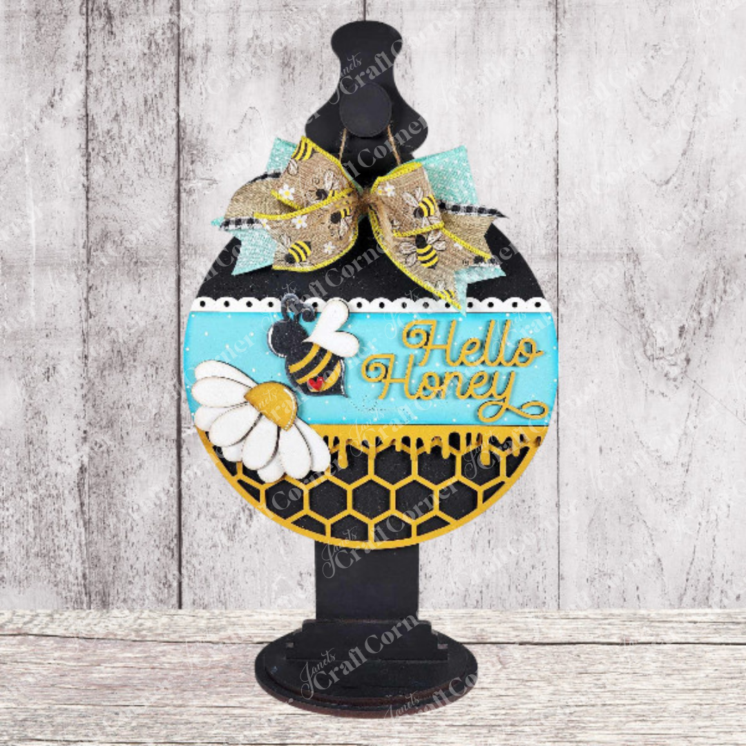 The "Hello Honey Wall Hanger" by Janet's Craft Corner is a DIY craft kit to create a decorative sign with a rounded silhouette on a stand, featuring bees, daisies, and "Hello Honey" in gold over honeycomb patterns, finished with a ribbon bow—ideal for rustic wooden home decor.