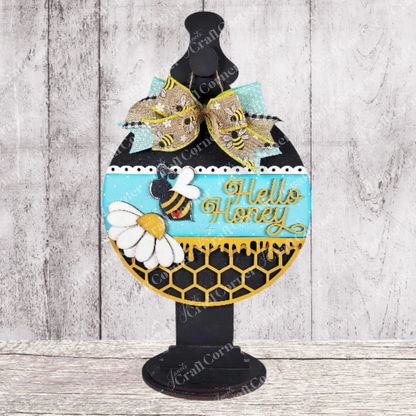The "Hello Honey Wall Hanger" by Janet's Craft Corner is a DIY craft kit to create a decorative sign with a rounded silhouette on a stand, featuring bees, daisies, and "Hello Honey" in gold over honeycomb patterns, finished with a ribbon bow—ideal for rustic wooden home decor.