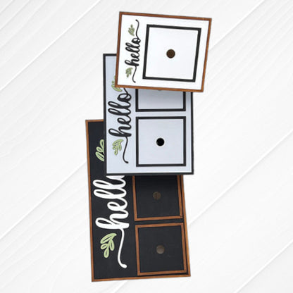 Presenting the Hello Interchangeable Squares Holder from Janet's Craft Corner – a DIY Craft Kit featuring a rectangular signs adorned with a design, showcasing the word "hello" and elegant decorative leaves. This distinctive home décor piece includes a central round hole for effortless changing of the squares, beautifully displayed on a white wooden surface.