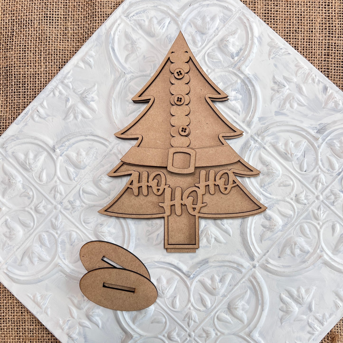 The Standing Christmas Tree Shelf Sitters from Janet's Craft Corner are charming wooden ornaments shaped like a Christmas tree, featuring cut-out details such as buttons, a belt, and "HO HO HO" letters. Ideal for holiday decor, these pieces are perfect for displaying on a textured white tile background and come with an accompanying oval wooden piece for added charm.